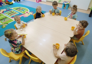 dzieci siedzą przy stoliku jedzą chrupki maczając je w kubku z miodem