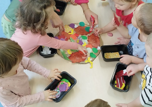 dzieci wraz z opiekunką układają na papierowej pizzy dodatki