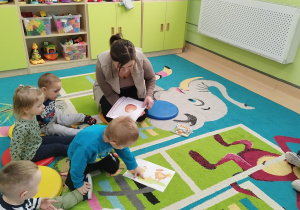dzieci siedzą na dywanie , opiekunka pokazuje im ilustracje
