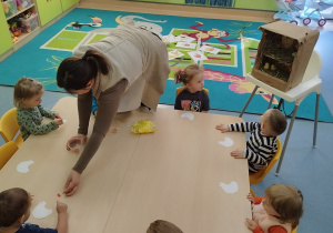 dzieci siedzą przy stoliku ,a opiekunka rozdaje im piórka