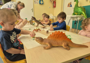 dzieci siedzą przy stolikach, na których stoją figurki dinozaurów, i za pomocą gąbki malują gipsowe dinozaury