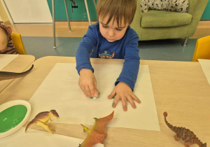 chłopiec siedzi przy stoliku i za pomocą gąbki pokrywa zieloną farbą gispową figurkę dinozaura