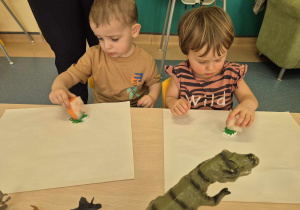 dzieci siedzą przy stoliku i malują gąbką maczaną w farbie gipsowe dinozaury