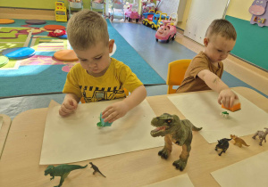 chłopcy siedzą przy stoliku i za pomocą gąbki maczanej w zielonej farbie malują gipsowe figurki dinozaurów