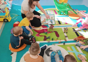 dzieci siedzą na dywanie , a opiekunka pokazuje im ilustracje z dinozaurami w książce