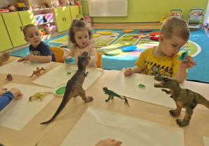 dzieci siedzą przy stoliku i malują gąbkami z farbą gipsowe dinozaury