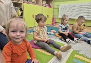 dzieci siedzą na dywanie, a dziewczynka uśmiecha się