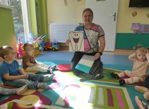 dzieci siedzą na dywanie, opiekunka pokazuje ilustracje uśmiechniętego zęba