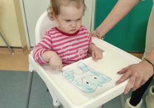 dziewczynka siedzi w krześle, przed nią karta z wykonanym zadaniem plastycznym