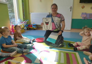 opiekunka pokazuje ilustrację wesołego zęba dzieciom siedzącym na dywanie