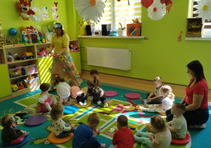 dzieci siedzą na dywanie wraz z opiekunką, jedna chodzi i rozrzuca kwiaty