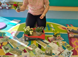 opiekunka pokazuje kwiaty w doniczkach, na dywanie leżą zdjęcia i ilustracje kwiatów i zwierząt