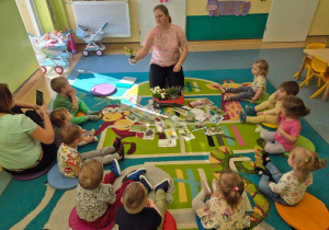 dzieci siedza na dywanie, opiekunka pokazuje wiosenne kwiaty w doniczkach