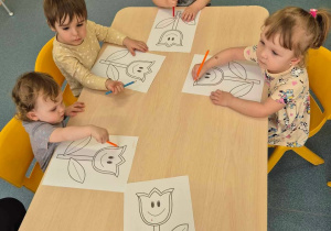 dzieci siedzą przy stoliku z kredkami w ręce, przed nimi kartki z wydrukowanym tulipanem