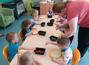 dzieci siedzą przy stoliku, opiekunka pomaga chłopcu, tłumaczy jak wykonać pracę