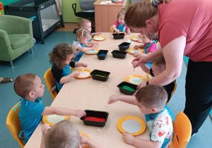 dzieci siedzą przy stoliku, z farbami i talerzykami z mlekiem, opiekunka pomaga chłopcu , tłumaczy jak wykonać pracę