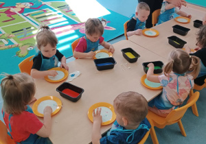 dzieci siedzą przy stoliku, opiekunka pomaga chłopcu wykonać prace