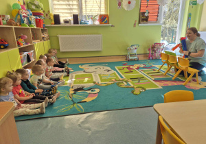 dzieci siedzą na dywanie, opiekunka przed nimi prezentuje mini teatrzyk
