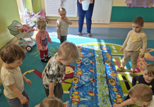 opiekunka stoi i trzyma wiaderko, a dzieci na dywanie zbierają papierowe rybki