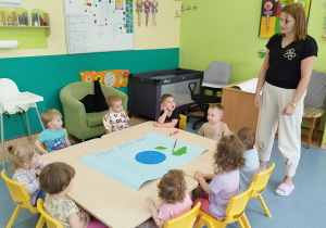 opiekunka stoi i opowiada siedzącym, przy stoliku dzieciom