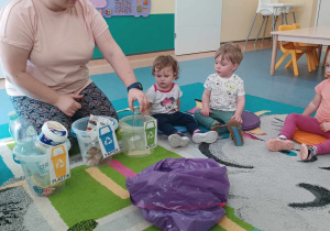 opiekunka siedzi z dziećmi na dywanie, przed nią stoją wiaderka i leży worek ze śmieciami