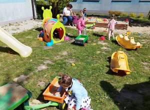 opiekunka z grupką dzieci siedzi w piaskownicy, a na pierwszym planie dziewczynka wysypuje piasek z przyczepki