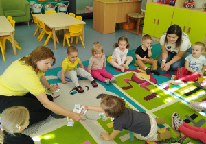 dziewczynka podaje opiekunce obrazek, wszyscy siedzą na dywanie i patrzą