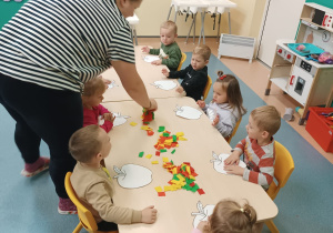 opiekunka pomaga dzieciom siedzącym przy stoliku w klejeniu bibuły