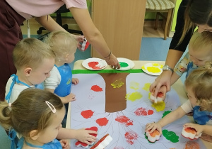dzieci stemplują na kartonie liście, opiekunki patrzą i pomagają