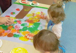 dzieci stoją wokół stolika, na którym leży karton z namalowanym już przez nie drzewem