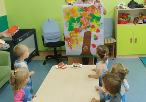 opiekunka pokazuje dzieciom stojącym przy stoliku karton z namalowanym przez nie drzewem