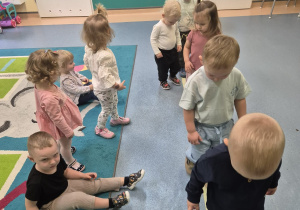 dzieci idą jedno za drugim, niektóre dzieci siedzą na dywanie,opiekunka pomaga