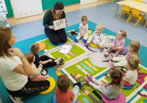 dzieci i opiekunki siedzą na dywanie, opiekunka pokazuje ilustracje