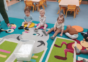 dzieci siedza na dywanie, a przed nimi stoi pojemnik ze śniegiem