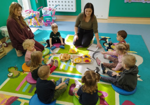 dzieci i opiekunki siedzą na dywanie, mówią co ciocia przyniosła na tacy