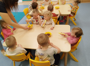 dzieci siedzą przy stoliku, trzymają chrupki, opiekunka podaje miseczkę z czekoladą