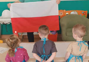 opiekunka pokazuje dzieciom plagę Polski, dzieci w fartuszkach stoją prze stoliku
