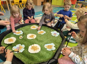 dzieci siedzą wokół pola , które jest łąką i nawlekają kółeczka na druciki