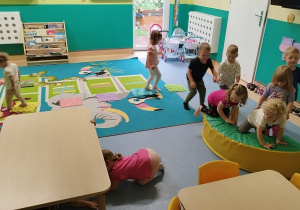 dzieci chodzą jedno za drugim pokonując przeszkodę, przechodząc pod stolikiem i depcząc płytki sensoryczne