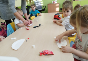 dziewczynka przykleja oczko do buzi Mikołaja, w tle dzieci i opiekunka pomagająca przyklejać elementy