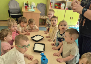 dzieci stoja przy stoliku, naszykowane pojemniki, konewka i nasionka