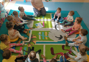 dzieci siedzą na dywanie , a opiekunka pokazuje planszę