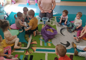 dzieci siedzą na dywanie z opiekunką, leży worek ze śmieciami i stoją wiaderka na śmieci