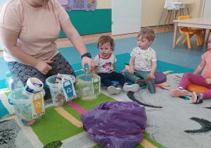 opiekunka siedzi z dziećmi na dywanie, przed nią stoją wiaderka i leży worek ze śmieciami