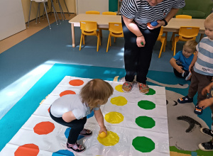 na dywanie w kolorowe kropki dziewczynka kuca z żółtą piłeczką, a opiekunka patrzy