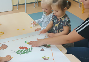 chłopiec stoi patrzy, a dziewczynka wraz z opiekunką nakleja