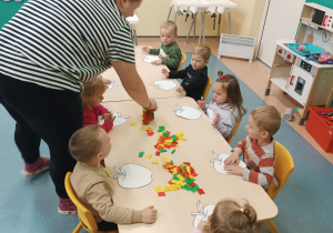 opiekunka pomaga dzieciom siedzącym przy stoliku w klejeniu bibuły