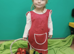 dziewczynka w fartuszku trzyma w ręce koszyk z jabłkami