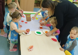 dzieci przy stoliku maczają gąbki w farbie i stemplują liście na kartonie , opiekunki pomagają