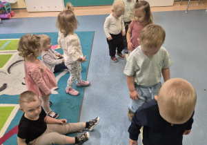 dzieci idą jedno za drugim, niektóre dzieci siedzą na dywanie,opiekunka pomaga
